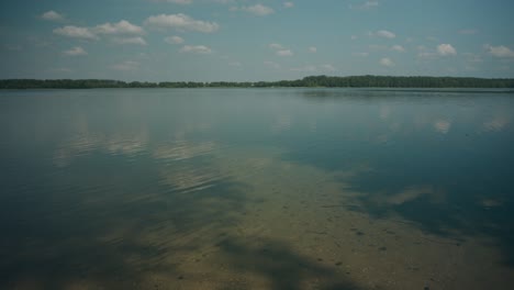 Calm-lake-waters-reflect-sparse-cloudy-sky-to-treeline-horizon