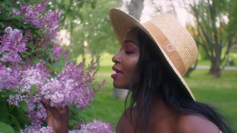 Black-woman-in-summer-dress-and-straw-hat-smells-pink-Lilac-flowers-in-park,-smiles-at-pleasant-smell---close-up-slow-motion