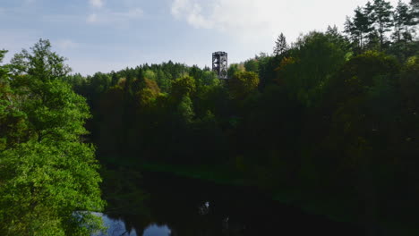 Beautiful-drone-view-flying-above-the-river-through-treetop-walkway-tower-with-people-on-it-between-green-woods-located-in-Anyksciai,-Lithuania,-eastern-Europe