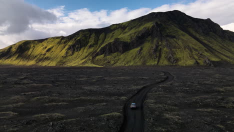 Aerial-push-in-shot-of-a-car-parking-on-a-black-volcanic-road-with-a-mountain-in-the-background,-on-a-sunny-day-with-some-clouds