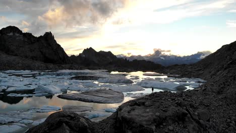 Aerial-flyover-above-a-lake-full-icebergs-from-a-melting-glacier-in-remote-parts-of-the-Swiss-Alps-with-a-hiker-enjoying-the-sunset-glow-and-view