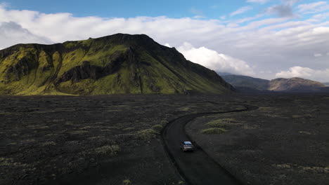 Aerial-push-in-shot-following-a-car-on-a-black-volcanic-road-with-a-mountain-in-the-background,-on-a-sunny-day-with-some-clouds