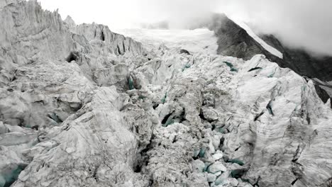 Aerial-flyover-through-the-crevasses-of-the-Moiry-glacier-near-Grimentz-in-Valais,-Switzerland-with-a-pan-down-view-from-the-mountain-peaks-to-the-ice-on-a-cloudy-summer-afternoon