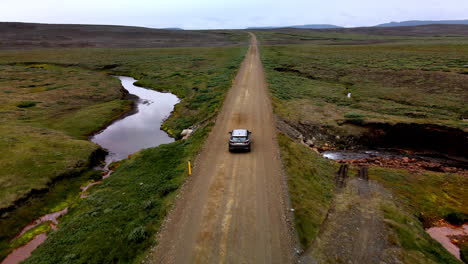 Aerial-tracking-shot-of-a-car-from-behind-on-a-lonely-mountain-gravel-road,-surrounded-by-green-bushes-in-Iceland