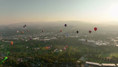 Ultra-wide-view-of-many-hot-air-balloons-soaring-through-the-air-above-a-vast-green-space