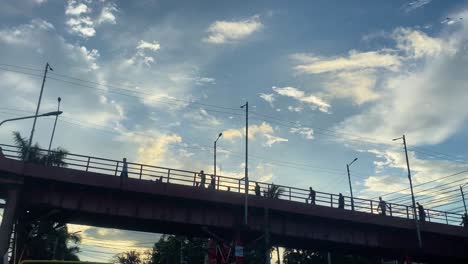 Looking-Up-At-Sillhouute-Of-People-Walking-Across-Bridge-In-Sylhet-With-Blue-Sky-And-Clouds-In-The-Background