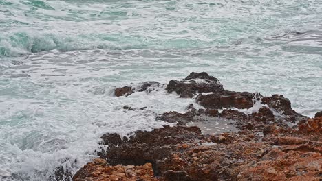 Coast-in-a-bad-weather-day,-gray-sky,-blue-waves-crashing-into-orange-rocks-of-the-coastline-spraying-foam-in-the-wind