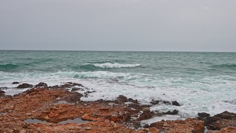 Sea-in-a-bad-weather-day,-gray-sky,-blue-waves-crashing-into-orange-rocks-of-the-coastline