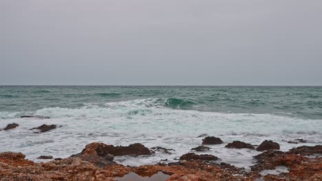 Ocean-in-a-bad-weather-day,-gray-sky,-blue-waves-crashing-into-orange-rocks-of-the-coastline