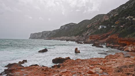 Coast-in-a-bad-weather-day,-gray-sky,-blue-waves-crashing-into-orange-rocks-of-the-coastline
