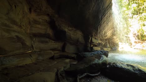 POV-inside-cave-looking-out-to-lagoon-and-waterfall-in-rainforest-on-sunny-day