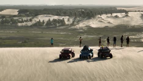 Aerial-orbiting-view-of-a-group-standing-on-the-edge-of-a-sand-dune-near-off-road-vehicles-overlooking-the-dramatic-views-as-strong-wind-blows-sand-across-the-face-of-the-dune
