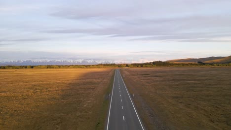 Straight-countryside-highway-leading-towards-a-stunning-snow-capped-mountain-range-during-a-golden-hour-in-Canterbury,-New-Zealand