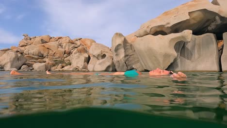 Half-underwater-scene-of-woman-in-blue-bikini-relaxing-floating-on-sea-water-of-Cala-Della-Chiesa-bay-with-eroded-granitic-rocks-in-background-of-Lavezzi-island-in-Corsica,-France