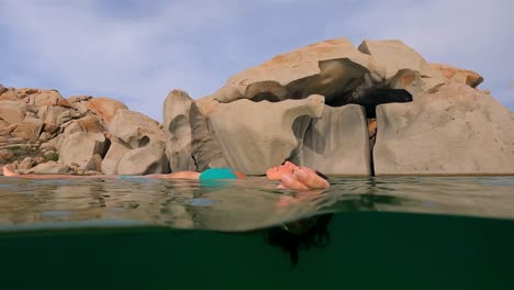 Half-underwater-scene-of-woman-in-blue-bikini-relaxing-floating-on-sea-water-of-Cala-Della-Chiesa-lagoon-with-eroded-granitic-rocks-in-background-of-Lavezzi-island-in-Corsica,-France