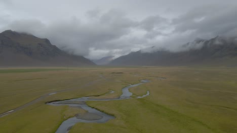 Icelandic-Landscape,-Drone-flyover-River-zigzagging-among-green-meadow,-Mountains-Background