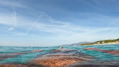 Personal-perspective-of-man-legs-and-feet-relaxing-while-floating-on-turquoise-sea-water-of-Saleccia-beach-and-lagoon-in-background-on-Corsica-island-in-France