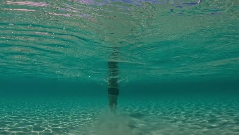 Underwater-back-view-in-shallow-water-of-man-walking-on-seabed-raising-clouds-of-sand-floating-in-turquoise-tropical-sea