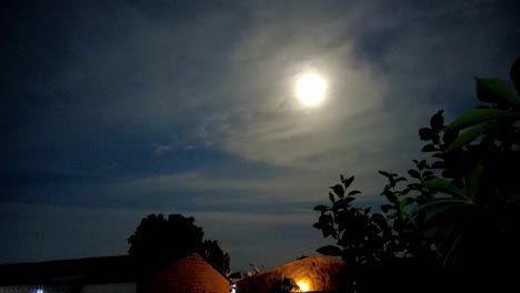 Moon-motion-lapse-from-left-to-right-with-visible-house-roofs-and-plants-on-slightly-cloudy-night