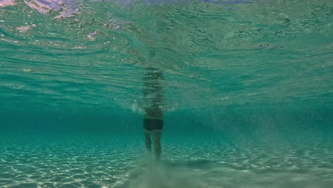 Underwater-back-view-in-shallow-water-of-man-walking-on-seabed-raising-clouds-of-sand-floating-in-turquoise-tropical-sea