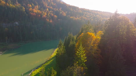 Orbiting-aerial-view-across-sunlit-emerald-Stausee-lake-glowing-autumn-alpine-woodland-valley