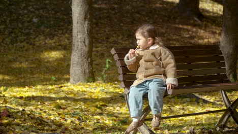 Cute-Little-Girl-Sitting-on-a-Banch-in-Autumn-Park-Enjoying-Sucking-Sweet-Lollipop-Candy-outdoors---full-body-in-Slow-motion