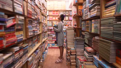 Asian-girl-standing-between-the-rows-of-bookshelves-and-exploring,-Side-angle-shot