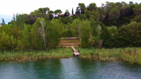 Flying-away-from-a-couple-sitting-in-a-wood-platform-in-front-of-a-lake-with-clear-watter,-between-the-trees-and-house-in-the-background,-in-Banyoles,-Catalonia-,-Spain