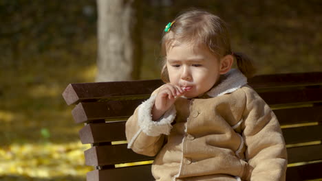Adorable-Blonde-Preschooler-Girl-Enjoy-Licking-Lollipop-Sitting-on-Banch-In-Autumn-Park-in-Slow-motion-at-sunset