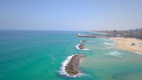 Sunny-Summer-Aerial-in-Israel,-Ashkelon-Beach---Pan-from-the-Ocean-over-rock-dams-to-the-city