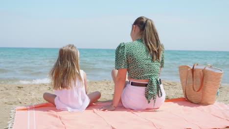 a-mother-and-daughter-sitting-back-to-back-on-an-orange-towel-in-the-sand-by-the-sea-on-a-sunny-day-with-their-hair-blowing-in-the-wind
