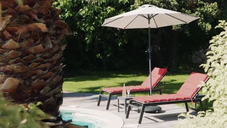 Zwei-Leere-Rote-Liegen-An-Einem-Pool-Mit-Weißem-Sonnenschirm-In-Einem-Luxuriösen-Garten-Mit-Palmenstamm-Und-Rasen