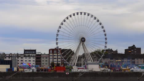 Slow-motion-reveal-shot-of-a-moving-Ferris-wheel-at-a-festival-in-cologne-with-other-rides-on-the-side-by-the-rhine-river-during-dusk