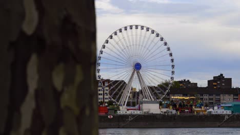 Slow-motion-reveal-shot-from-a-tree-trunk-to-a-moving-Ferris-wheel-at-a-folk-festival-in-cologne-in-germany-on-the-rhine-river-with-other-lighted-rides-during-dusk