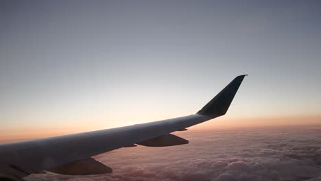 View-out-window-of-commercial-airline-plane-wing-with-soft-orange-and-magenta-hues-at-sunset-with-clouds-below