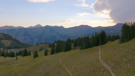 Aerial-flying-over-a-hiking-trail-and-tree-lined-ridge-and-towards-a-mountain-range-in-the-Colorado-Rockies-at-sunset