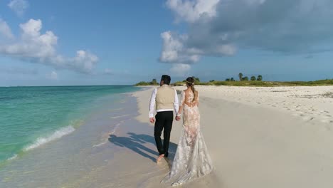 Couple-grown-and-bride-walking-holding-hands-on-white-sand-beach,-Man-pointing-sea-FROM-BEHIND