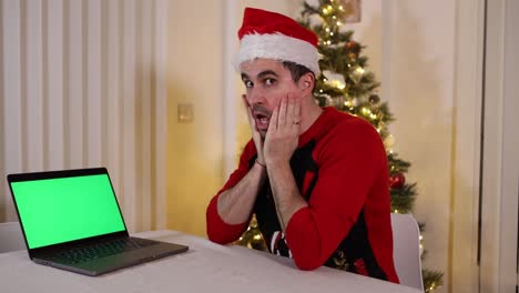 Festlicher-Mann-Mit-Weihnachtsmütze-Und-Pullover,-Der-Auf-Die-Kamera-Und-Den-Grünen-Bildschirm-Des-Laptops-Blickt,-Mit-überraschten-Schockausdrücken,-Die-Nicht-Glauben,-Was-Er-Gesehen-Hat