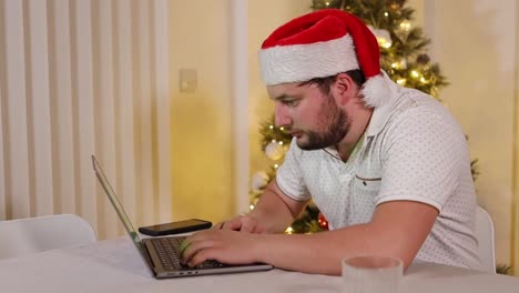Festive-bearded-male-wearing-Santa-hat-browsing-internet-on-laptop-computer-at-home-at-Christmas,-side-view