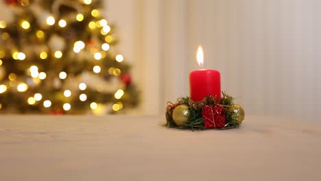 Einzelne-Rote-Festliche-Weihnachtskerzendekoration,-Die-Brennt-Und-Auf-Dem-Tisch-Sitzt,-Mit-Flachem-Fokus-Geschmückter-Weihnachtsbaum-Mit-Lichtern-Im-Hintergrund
