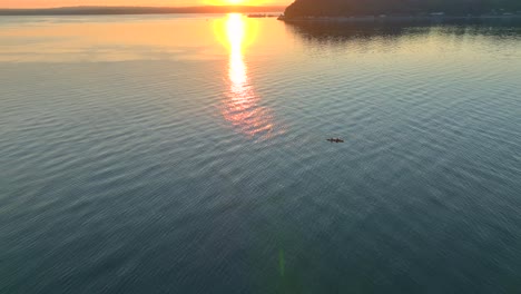 Aerial-drone-view-circling-left-of-two-people-in-a-sea-kayak-paddling-in-a-bay-near-Seattle-washington-at-sunrise-with-sun-glare-reflected-off-water