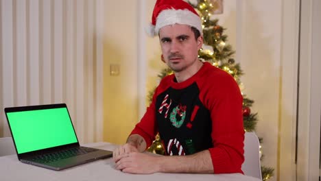 Male-wearing-Santa-hat-and-sweater-looking-at-camera-pointing-at-chroma-key-green-screen-laptop-with-unhappy-judgement-expressions