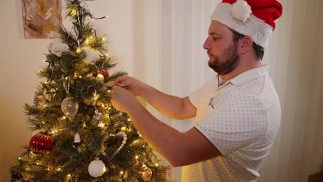 Hombre-Vestido-Con-Sombrero-Festivo-De-Santa-Decorando-El-árbol-De-Navidad-Tradicional-Arreglando-Luces-Y-Decoraciones-En-La-Noche