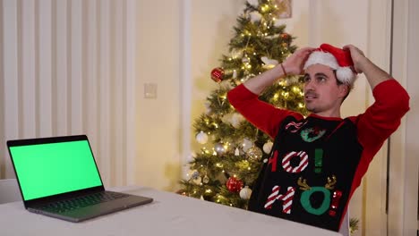 Kerl-Mit-Weihnachtsmütze-Und-Pullover,-Der-Sich-Auf-Einem-Stuhl-Mit-Einem-Laptop-Mit-Grünem-Bildschirm-Zurücklehnt,-Der-Sich-Zufrieden-Und-Glücklich-Fühlt-Und-Darüber-Nachdenkt,-Was-Er-Gesehen-Hat