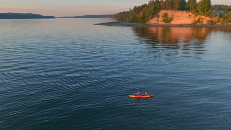 Aerial-drone-shot-circling-young-couple-sea-kayaking-paddling-in-bay-to-then-reveal-southworth-ferry-terminal-pier-near-seattle-washington-at-sunrise
