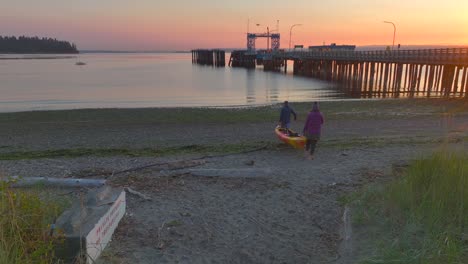 Man-and-woman-couple-walk-carrying-sea-kayak-out-to-the-ocean-bay-at-sunrise-near-Seattle-Washington
