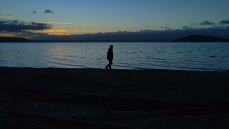 Lonely-Person-at-Dusk-slowly-walking-alone-along-shore-at-sunset-with-horizon-in-background