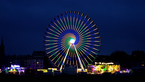 Static-handheld-shot-of-a-glowing-Ferris-wheel-in-different-colors-at-a-festival-in-cologne-in-germany-with-other-glowing-rides-and-attractions-at-night