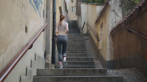 Slim-Caucasian-woman-running-and-jogging-upstairs-at-an-urban-city-alley-stairs,-still-shot-seen-from-behind