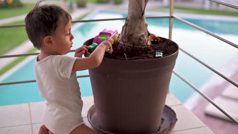 Young-latin-kid-playing-with-his-car-toys-on-the-dirt-inside-a-plantpot-at-his-home-balcony-watching-the-pool-downstairs